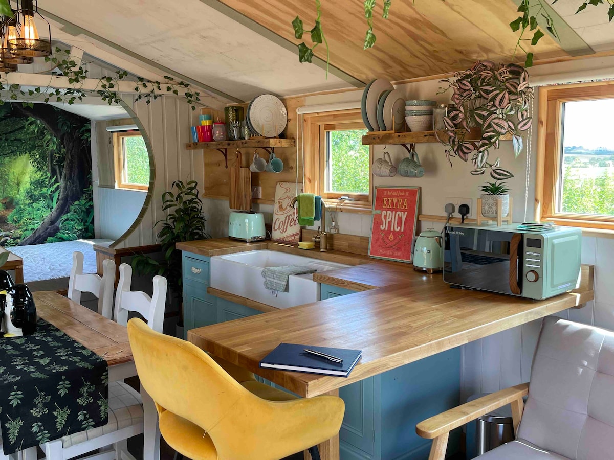 A cozy kitchen space is displayed with light wood cabinetry and a dining table. Bright green accents are found in decor and dishware. A microwave and kettle are visible along with various kitchen utensils. Natural light fills the room through two windows adorned with greenery.