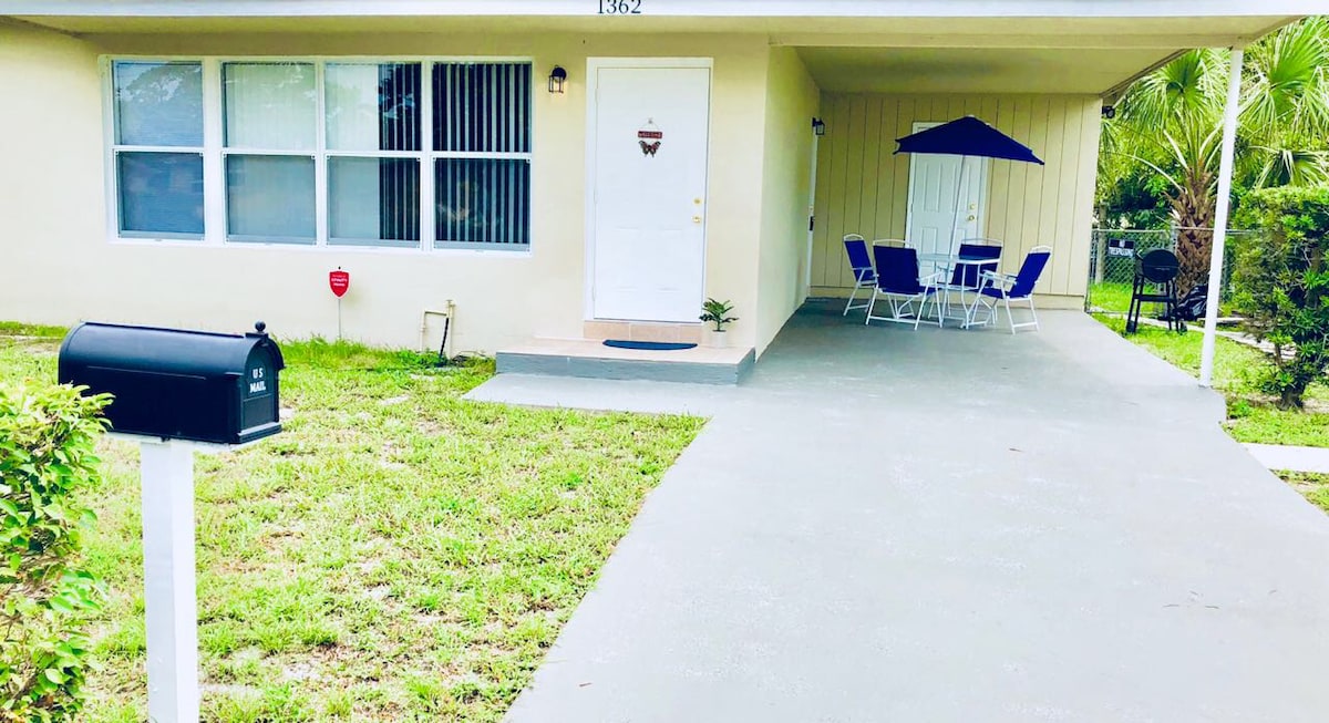 The exterior features a light-colored house with large windows allowing ample natural light. A welcoming front porch holds a door with a simple design. A small table and chairs in vibrant blue are positioned under a shade. A mailbox is visible near the entrance.