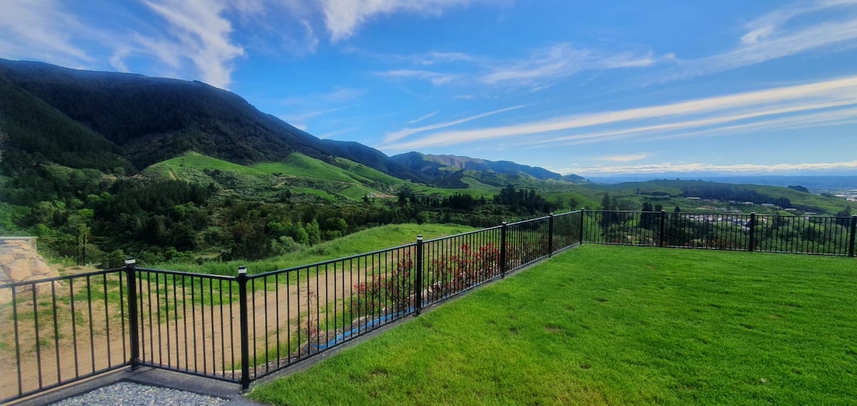 A scenic view of rolling green hills extends under a clear blue sky. A black metal railing encloses a grassy area, framing the vibrant landscape while providing an unobstructed sight of the mountains in the background.