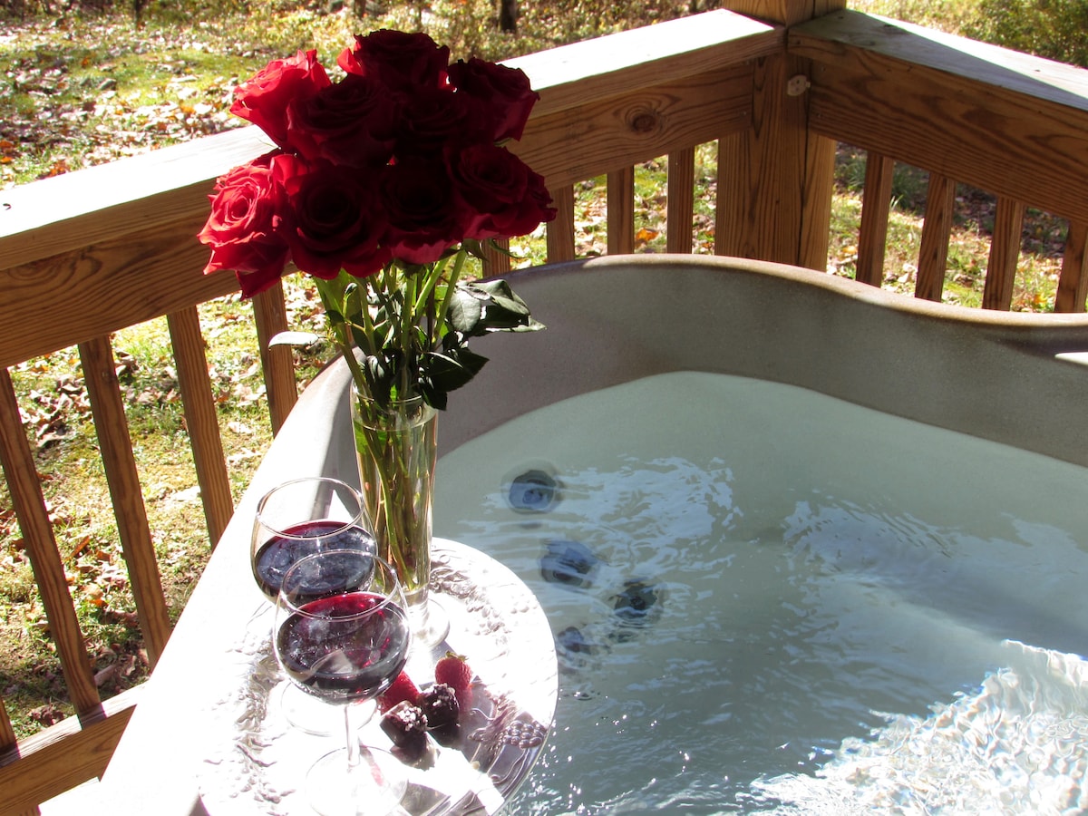 A hot tub is positioned on a wooden porch surrounded by nature. A bouquet of red roses stands tall in a vase. Two glasses filled with a dark beverage rest on the edge, accompanied by fresh berries.