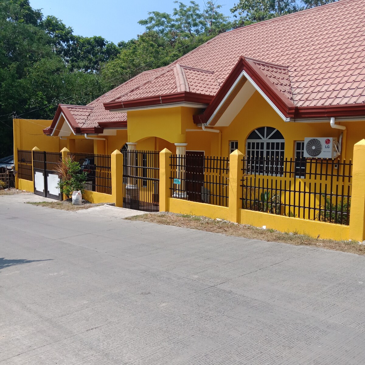 The exterior of a single-story home is depicted, featuring a bright yellow facade with a red roof. A gated entrance is visible, along with a well-maintained garden area. Nearby trees and greenery contribute to the surrounding environment. A wall-mounted air conditioning unit is positioned on one side.
