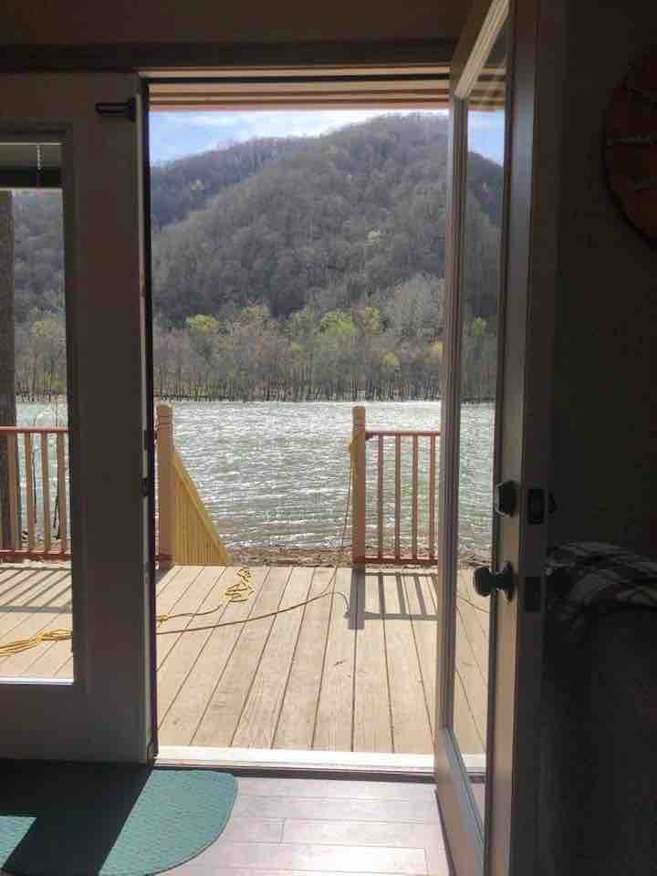 A door stands open, revealing a wooden deck that overlooks the tranquil New River. In the background, lush green mountains create a serene atmosphere. Sunlight filters through, casting a gentle glow on the wood surface and the water below.