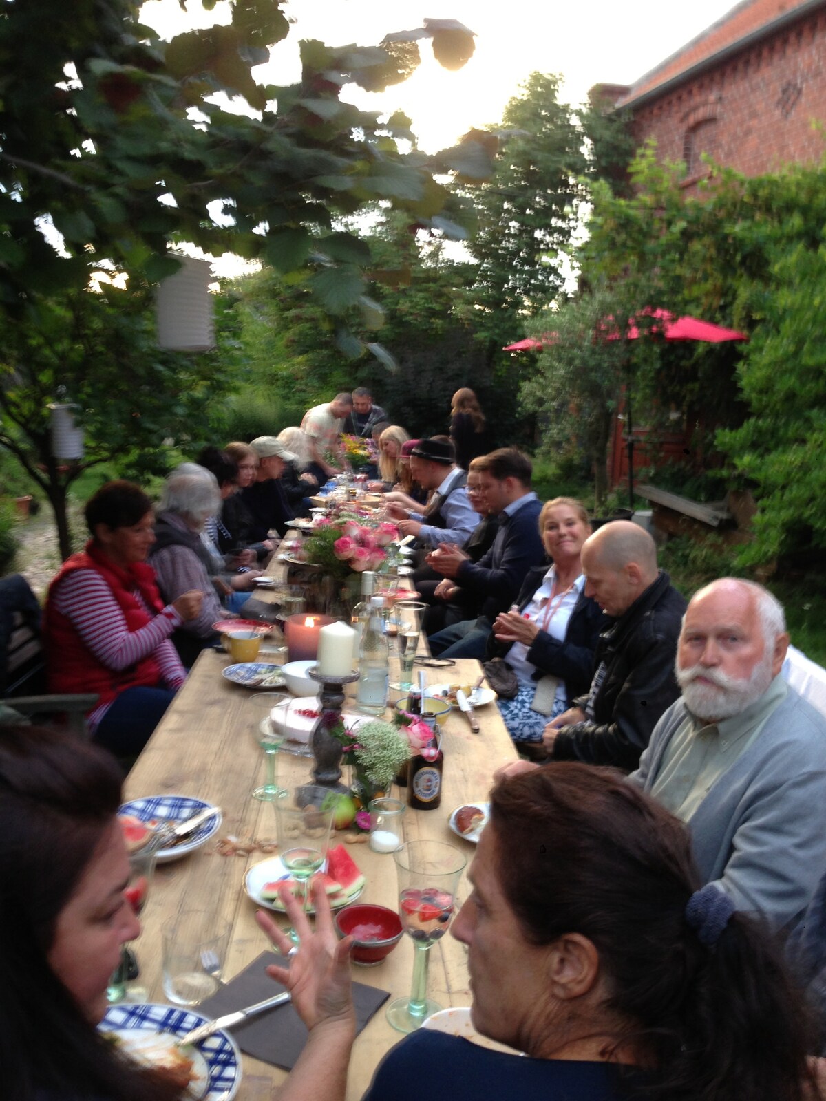 A long wooden table is set outdoors for a communal meal, surrounded by greenery and flowers. Guests are seen enjoying the gathering, with red umbrellas providing shade. Soft light during sunset creates an inviting atmosphere for socializing.