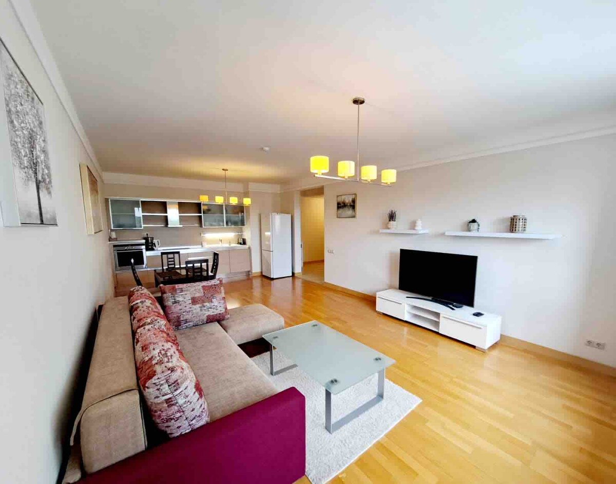 An open living area features a comfortable sectional sofa with decorative pillows in shades of purple and neutral tones. A glass coffee table sits in front, and a modern television is mounted on the wall. The kitchen is visible in the background, equipped with appliances and illuminated by pendant lighting.