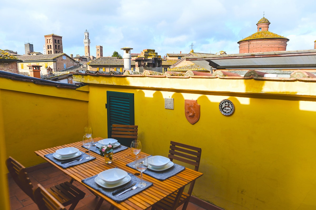 A sunlit terrace is presented, featuring a dining table set for four individuals. Wooden slatted chairs surround the table, while a backdrop of Siena's rooftops and historical towers can be admired. The bright yellow walls create a cheerful ambiance, enhancing the outdoor dining experience.