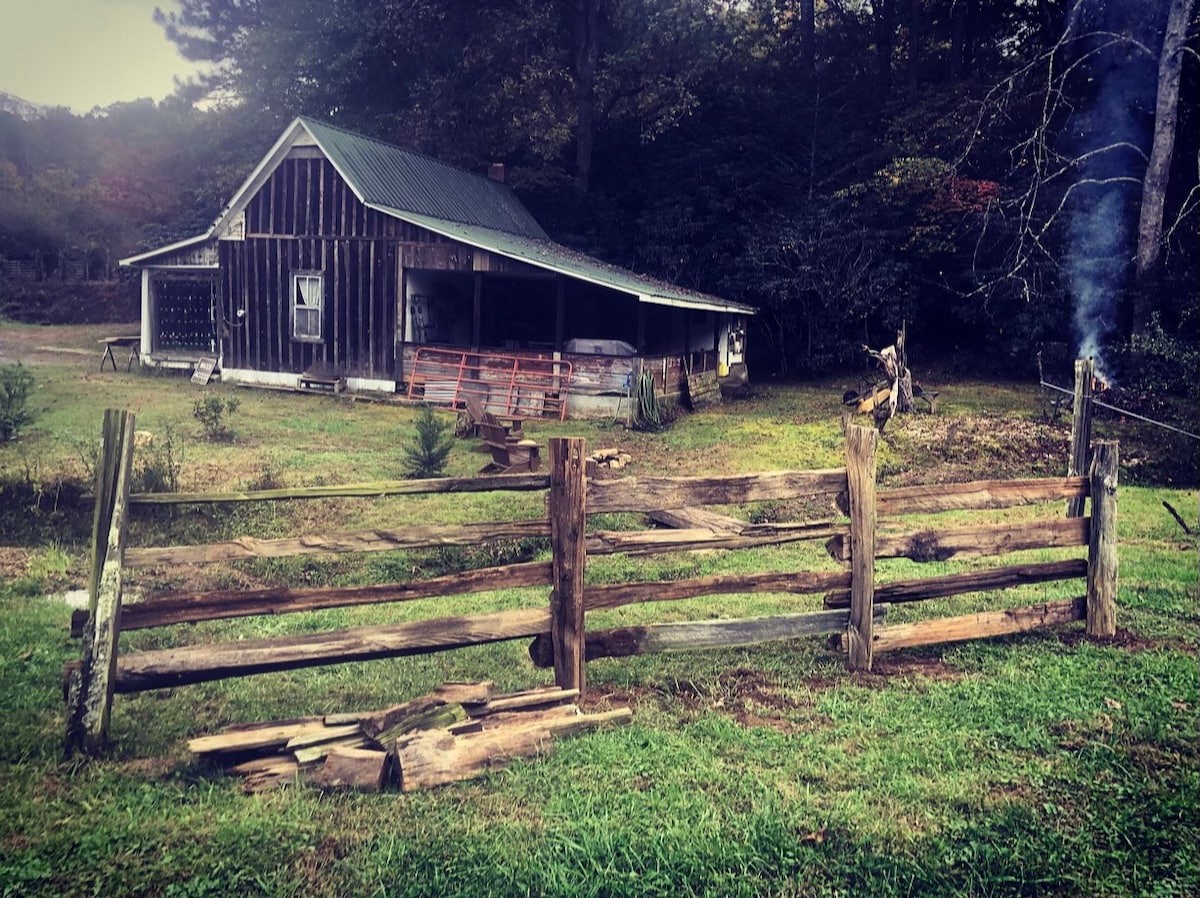 A rustic farmhouse with a weathered wooden exterior is surrounded by greenery. A natural wooden fence runs along the foreground, while gentle smoke rises from a nearby area. The structure features a green metal roof and a covered porch, enhancing the outdoor rustic charm.