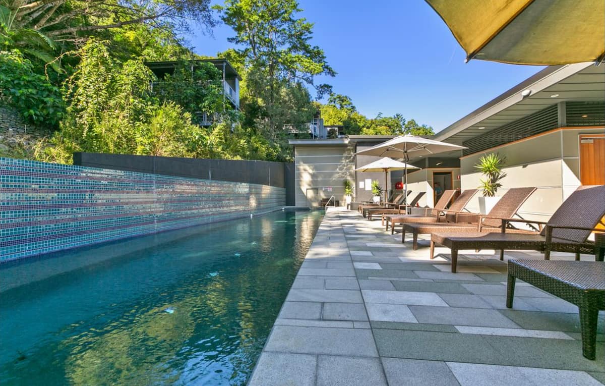 A serene pool area features a long lap pool bordered by stylish lounge chairs and large umbrellas. Lush greenery is visible in the background, enhancing the tranquil atmosphere. Stone tiles pave the surrounding space, offering a clean and contemporary look.