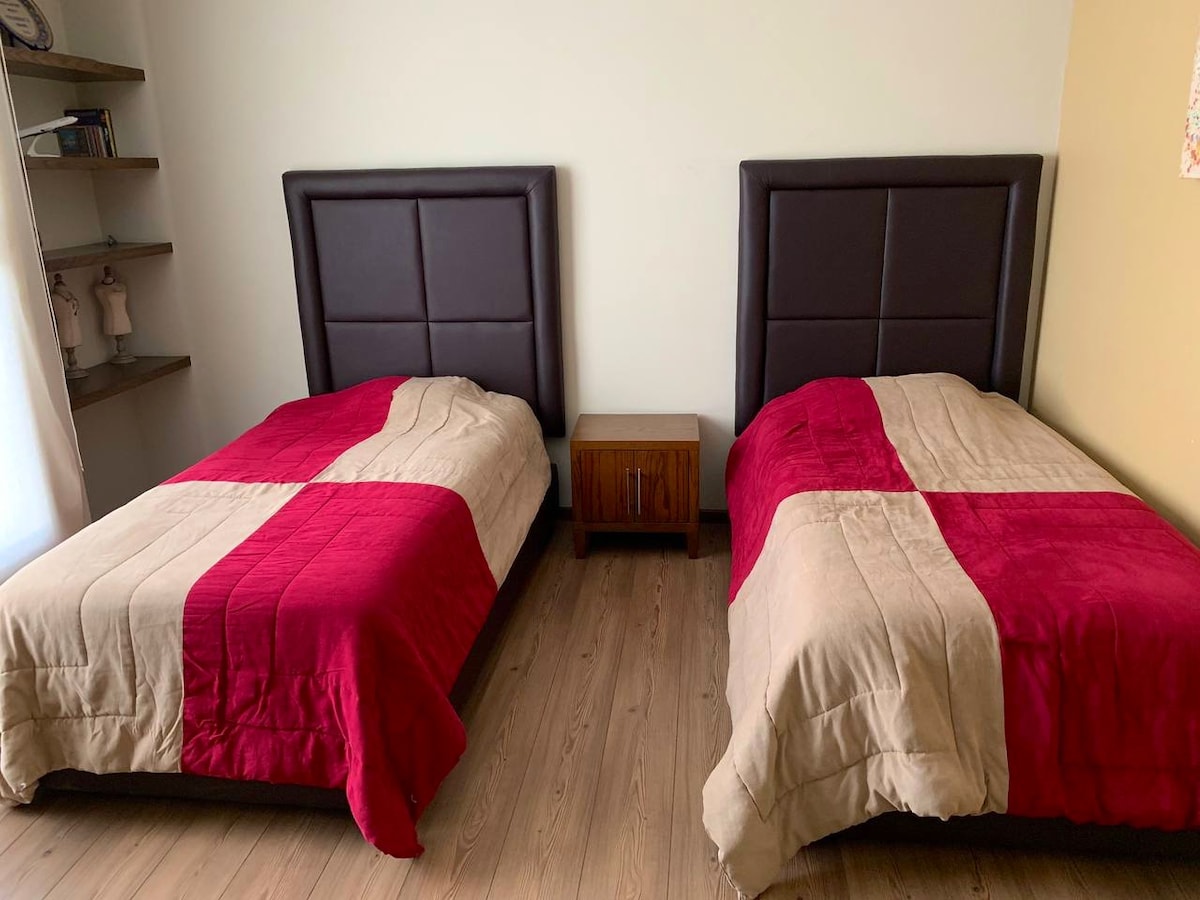 The image displays a room with two single beds positioned side by side, each covered with a red and beige quilt. A wooden nightstand is placed between the beds. The walls are painted in a light color, and a shelf with decorative items is visible in the background.