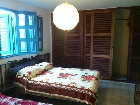 A spacious room is furnished with a large floral-patterned bedspread covering a bed. Two wooden shutters open to bright blue windows, allowing natural light to fill the space. A circular pendant light hangs above, and a wooden closet with slatted doors is positioned along one wall.