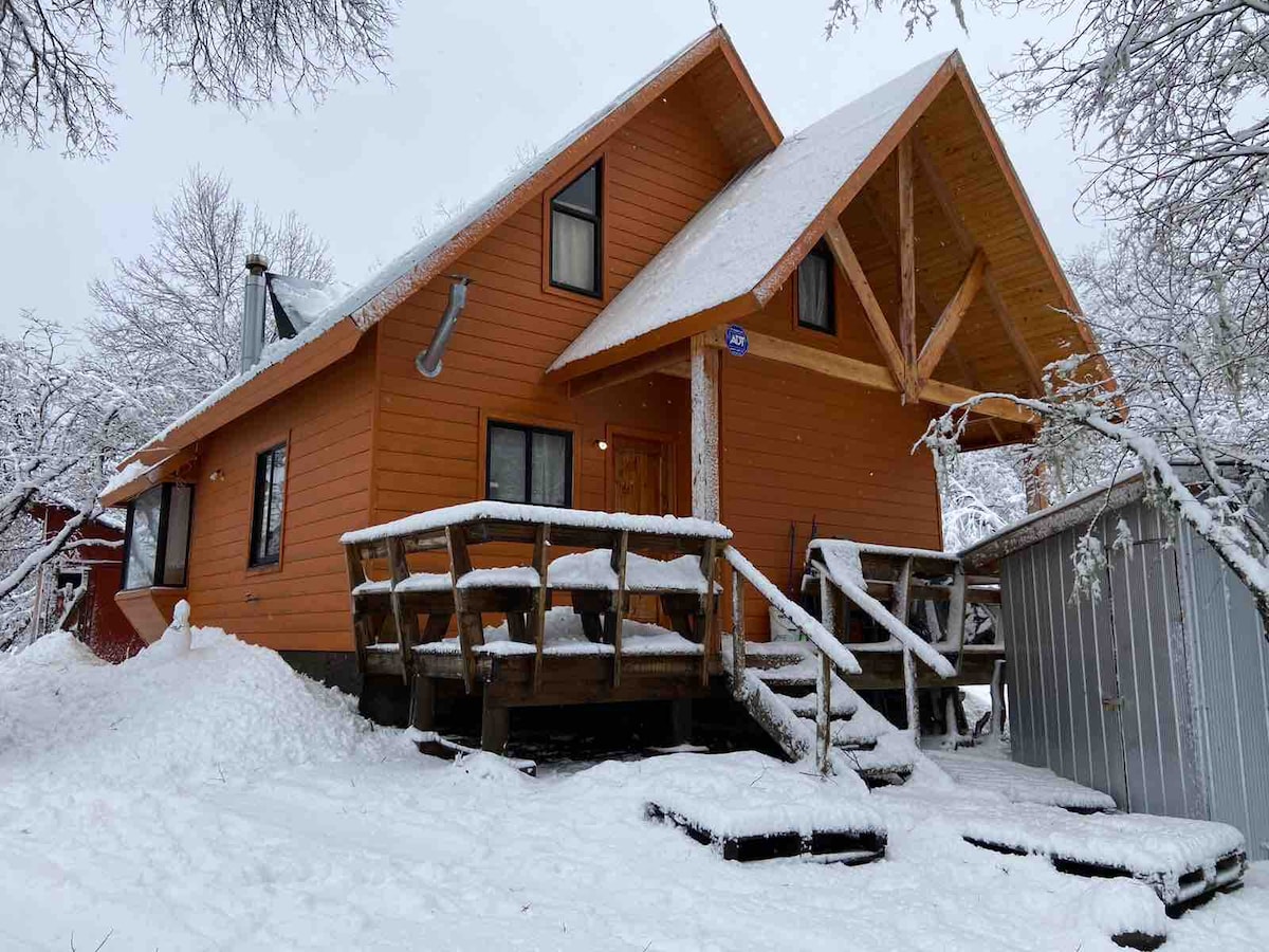 A two-story mountain cabin is set against a snowy landscape, featuring a warm orange exterior and a sloping roof. A wooden deck with railings surrounds the entrance, which is accentuated by steps leading up from a snow-covered ground.