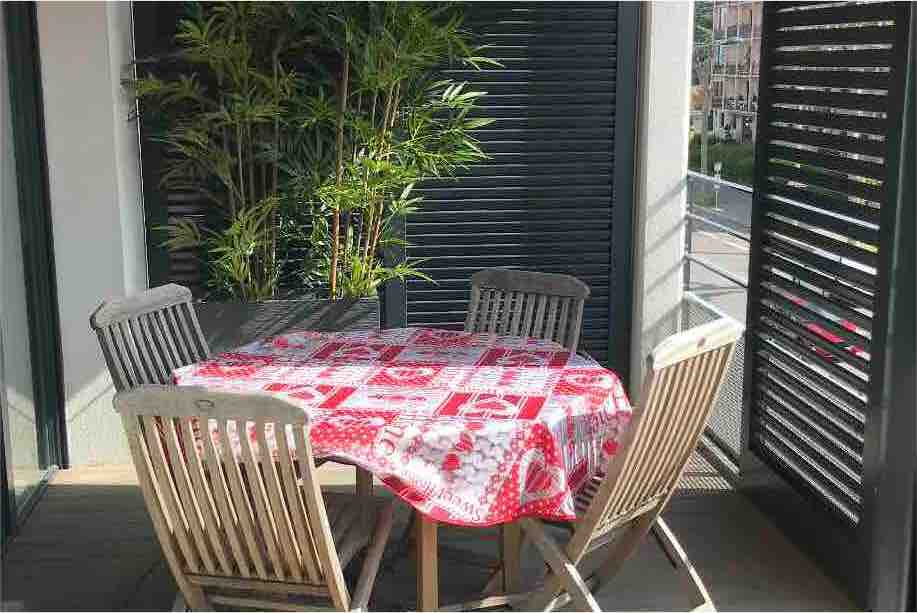 A terrace features a wooden table set with a red and white patterned tablecloth surrounded by four wooden chairs. Lush green plants add a natural element to the space, while the surrounding slatted shutters offer privacy and a sense of enclosure.