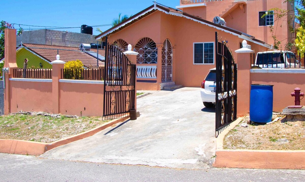 An inviting house exterior is showcased, featuring a gated entrance leading to a spacious driveway. The warm color of the house contrasts with the blue water barrel and the white vehicle parked nearby. Lush greenery is present along the sides of the property.
