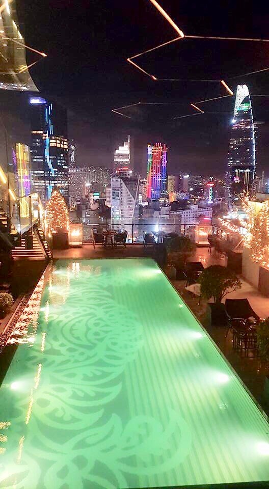 A rooftop pool is seen illuminated with elegant patterns, reflecting the night sky. The city skyline features vibrant, colorful lights from nearby buildings, creating a lively atmosphere. Seating areas are arranged around the pool, providing a space for relaxation and enjoyment of the view.