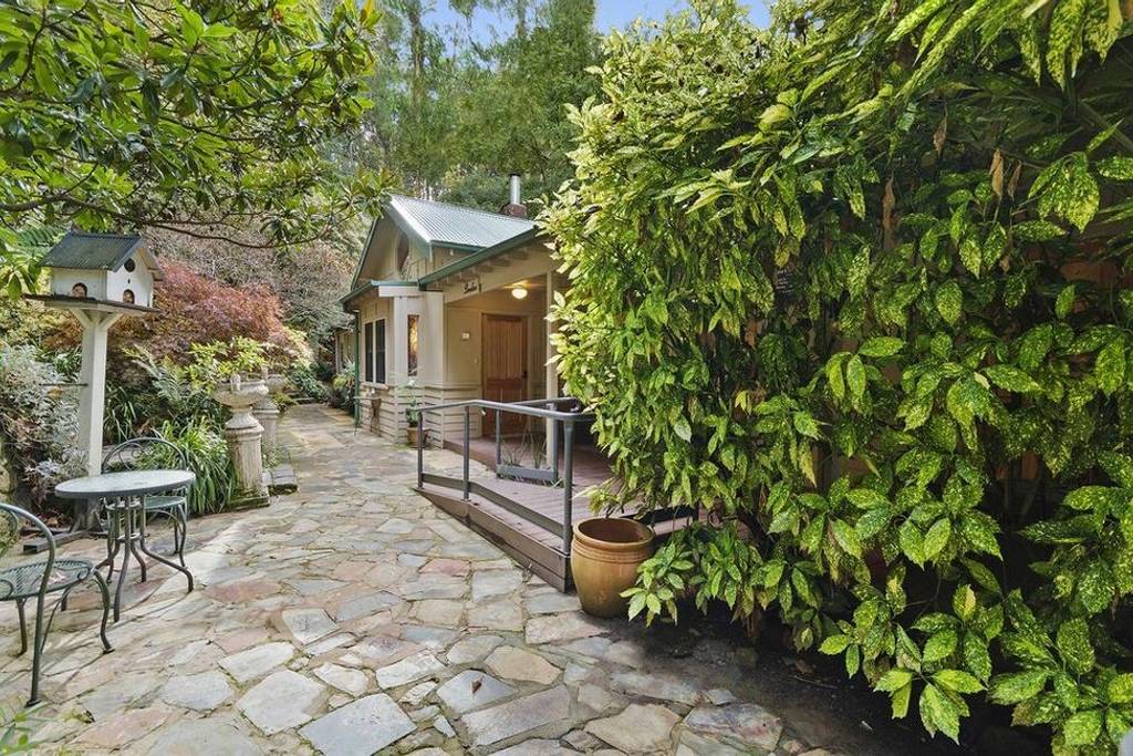 A stone pathway is bordered by lush greenery and vibrant foliage, leading to the entrance of a retreat. Outdoor seating with a small table and chairs is positioned nearby, creating a serene atmosphere surrounded by nature.