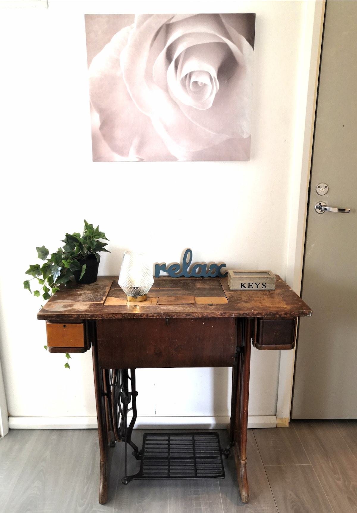 A vintage wooden desk is positioned against a wall, featuring two small drawers and a decorative black metal base. On the desk, a potted plant, a candle holder, and a sign displaying 'relax' and 'KEYS' are arranged. Above, a monochrome rose image is displayed.