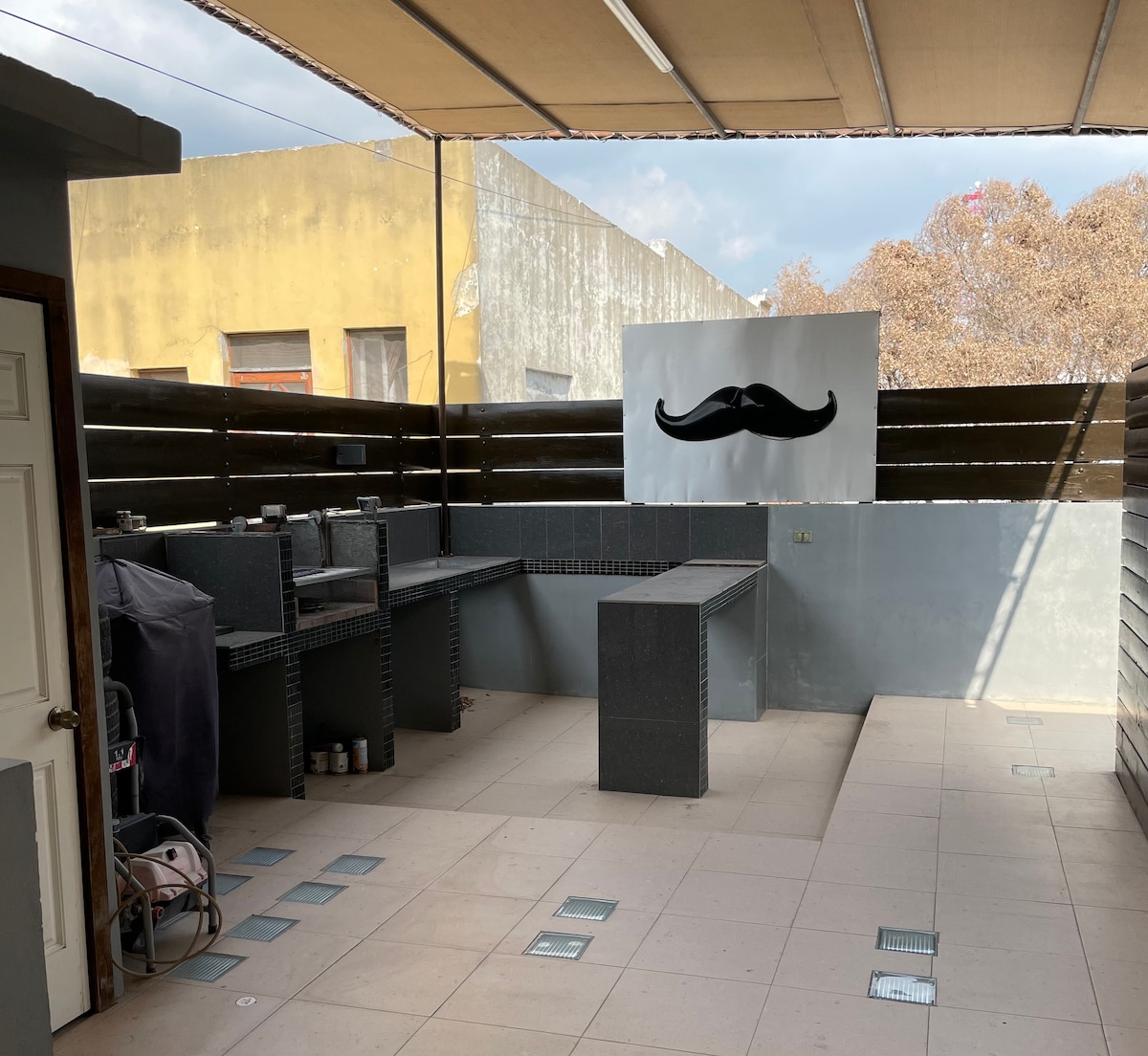 An outdoor terrace area is displayed, featuring a built-in grill and ample counter space for food preparation. The floor is tiled in neutral tones, and a mural of a mustache adds a whimsical touch to the wall. The space is partially shaded by an overhead awning.