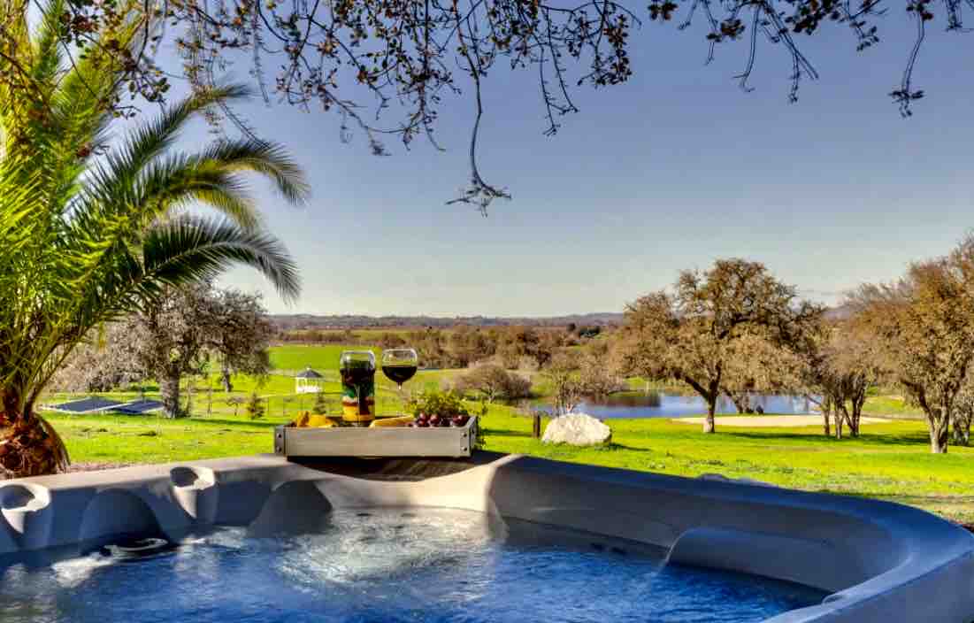 A private hot tub is set against a backdrop of lush greenery and a serene lake. Two glasses and a small platter with fruit are placed on the rim, complementing the tranquil atmosphere created by palm trees and distant rolling hills.