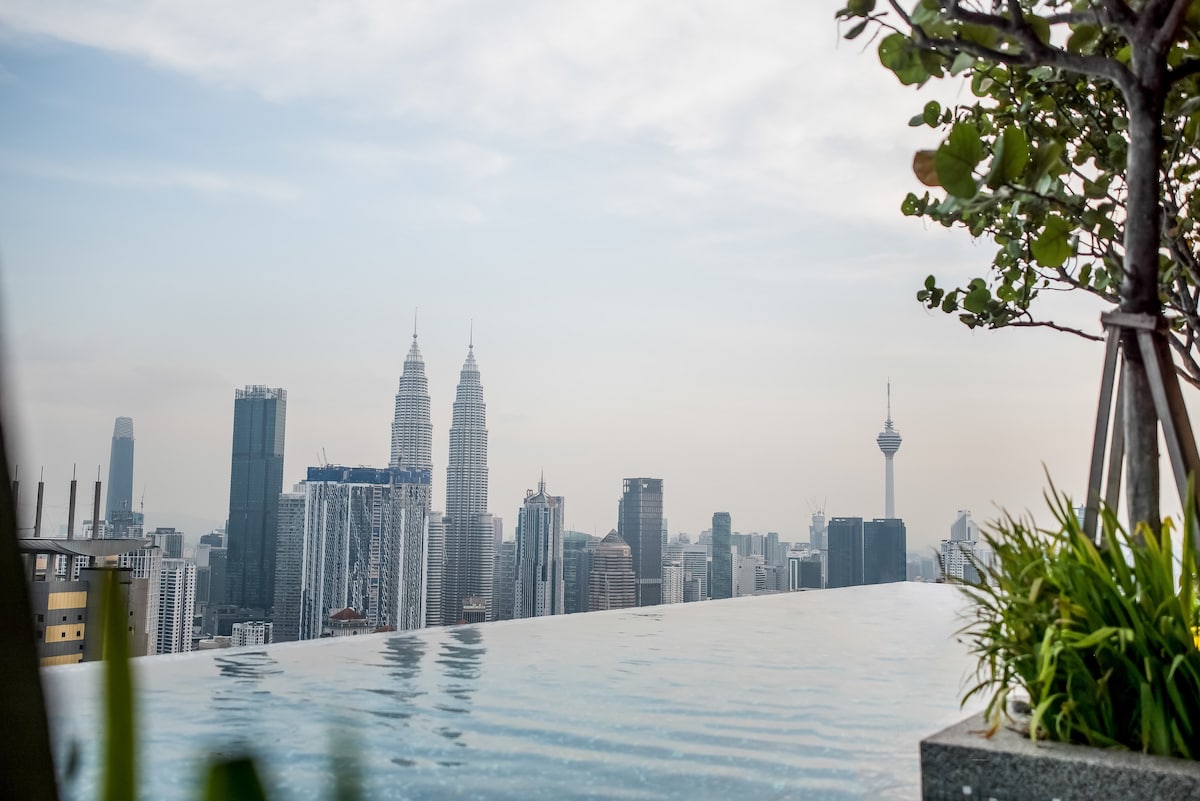 An infinity pool extends toward a panoramic city view, capturing iconic skyscrapers like the Petronas Twin Towers and the Kuala Lumpur Tower. Lush green plants frame the edge of the pool, enhancing the serene atmosphere of the rooftop space.
