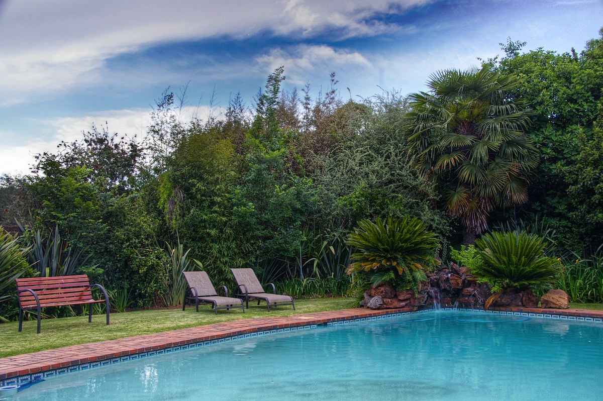 A serene pool area is framed by lush greenery and palm trees. Two lounge chairs are positioned beside the clear water, inviting relaxation. A small waterfall adds a calming element, while the surrounding landscape fosters a private retreat feel.