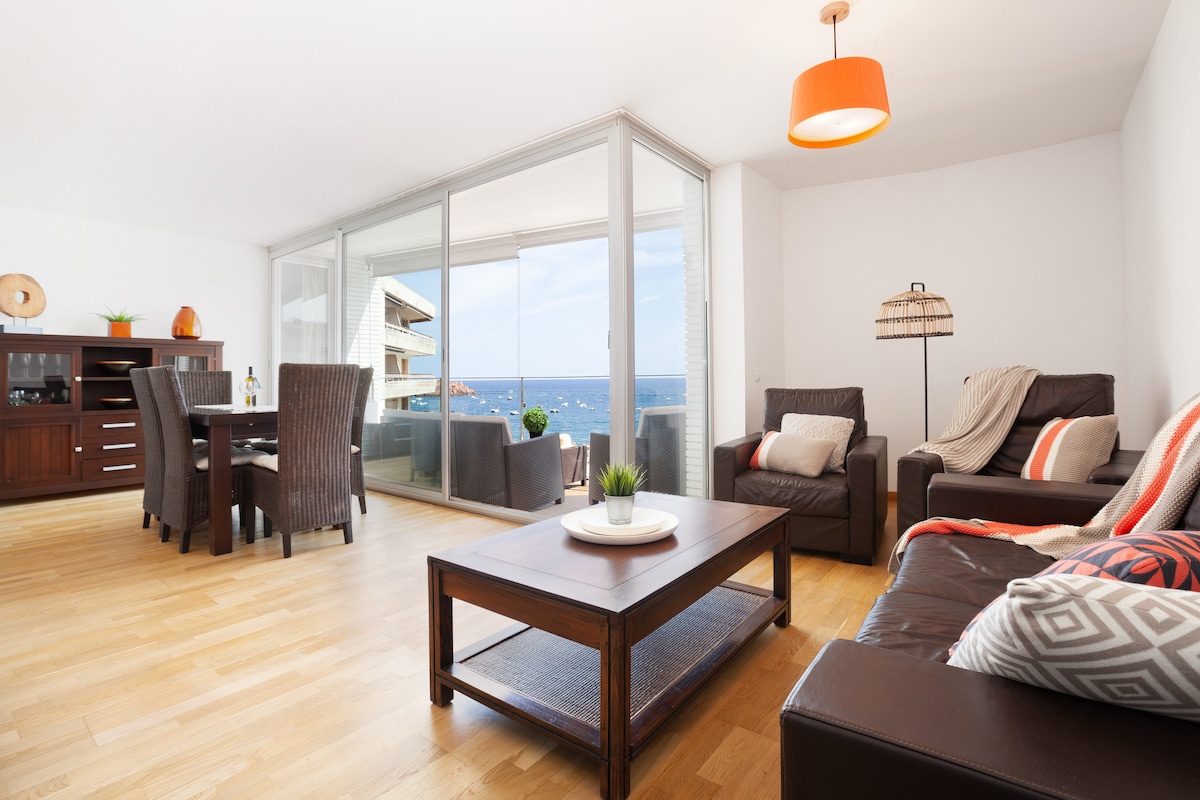 A spacious living area is highlighted by large glass doors offering sea views. Comfortable seating, including a leather sofa and armchairs, is arranged around a wooden coffee table. A dining table for four is positioned nearby, and natural light brightens the wood flooring.