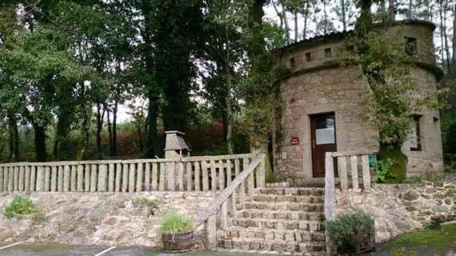 A circular stone structure is set within a wooded area, featuring a staircase leading up to a wooden door. Surrounding trees provide greenery, while a stone railing adds a rustic touch to the outdoor space.