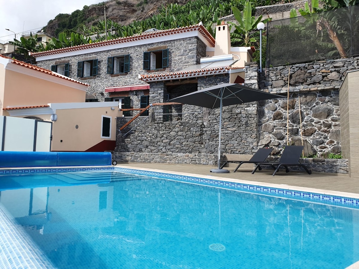 A stone house with traditional architecture is framed by lush banana trees. In the foreground, a spacious pool reflects the sky, accompanied by two sun loungers under a shaded umbrella. The surrounding terrace is paved, creating an inviting outdoor space for relaxation.