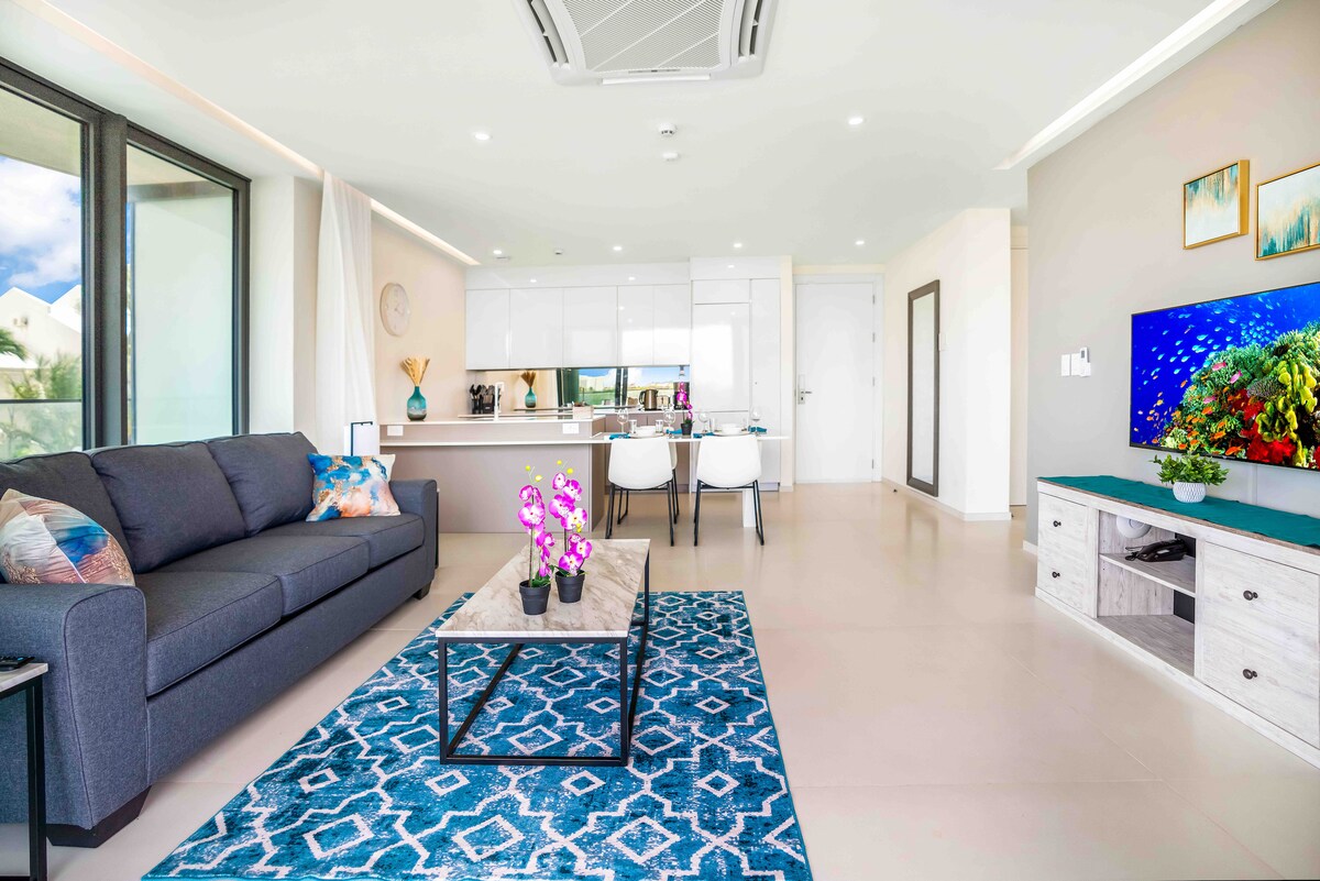 A modern living room features a dark grey sofa adorned with colorful cushions. A sleek coffee table sits on a blue patterned rug, accompanied by a potted orchid. Open kitchen cabinetry is visible in the background, complemented by a bright, airy atmosphere from large windows.