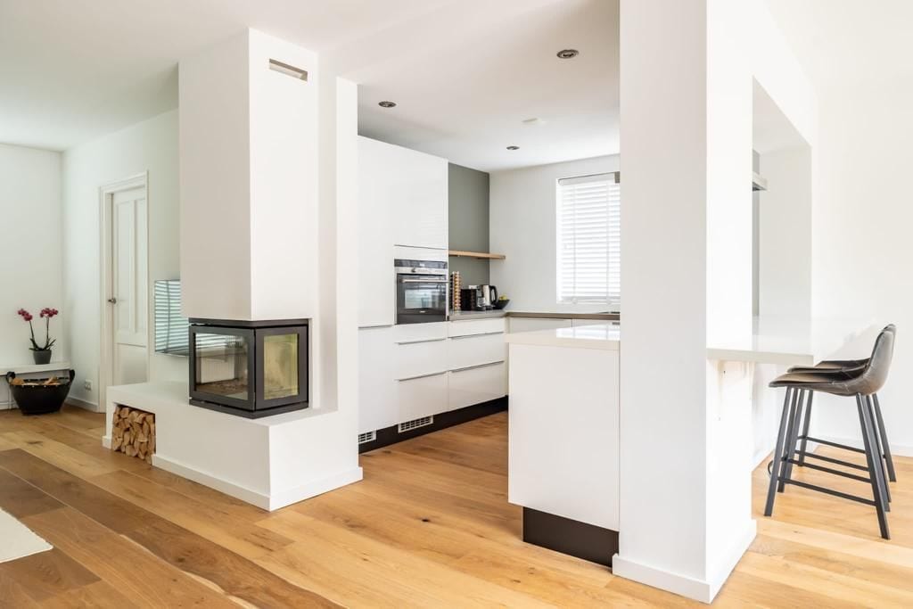 An open-plan kitchen area features a modern design with white cabinetry and a sleek chimney. Natural light fills the space through a window, highlighting the warm wooden flooring. A bar with high stools and a cozy fireplace are noticeable in this inviting setting.