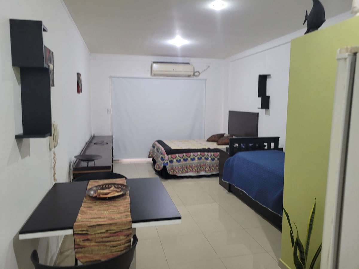 A modern room features a spacious bed and a second bed, both with decorative bedding. A television is mounted on the wall, and a small dining table is set with tableware. Natural light enters through a large window, enhancing the light-colored walls.
