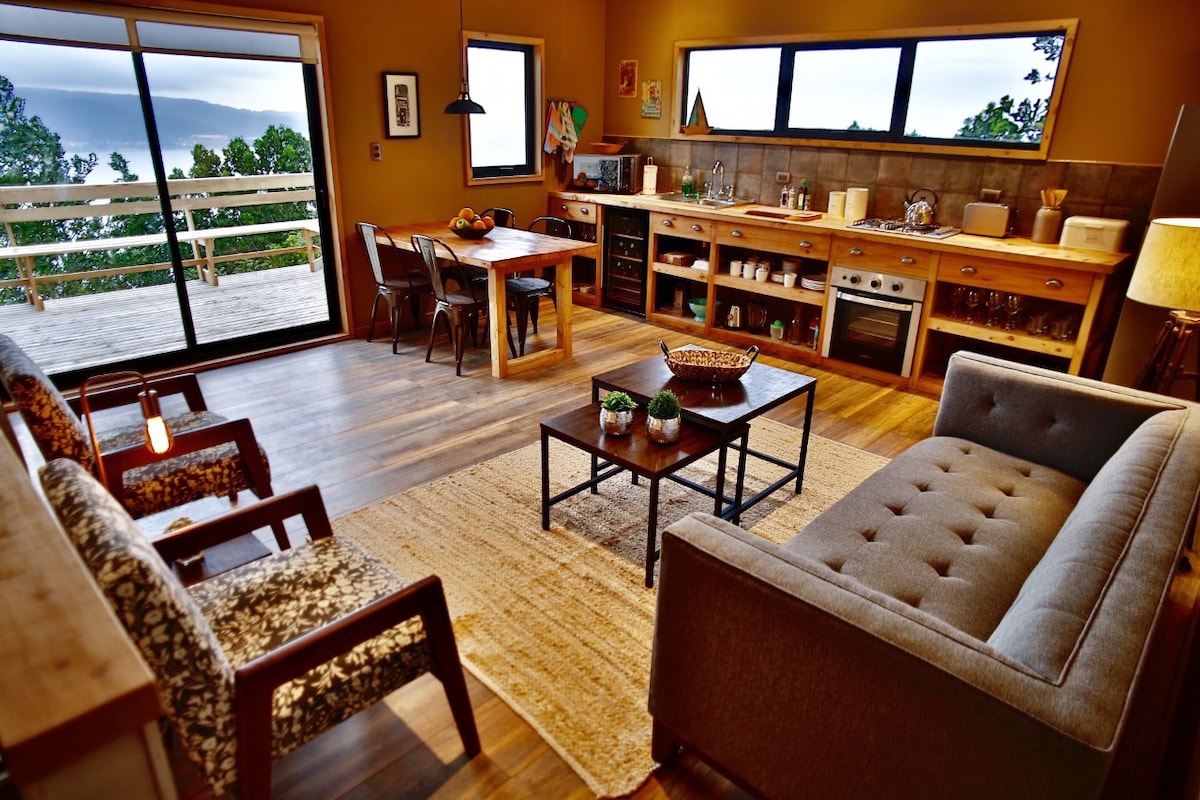 An open-concept living area showcases a cozy seating arrangement with a gray sofa and patterned chairs. A wooden dining table accommodates four. Large windows offer expansive views of the outdoor terrace and surrounding landscape, while warm lighting enhances the welcoming atmosphere.