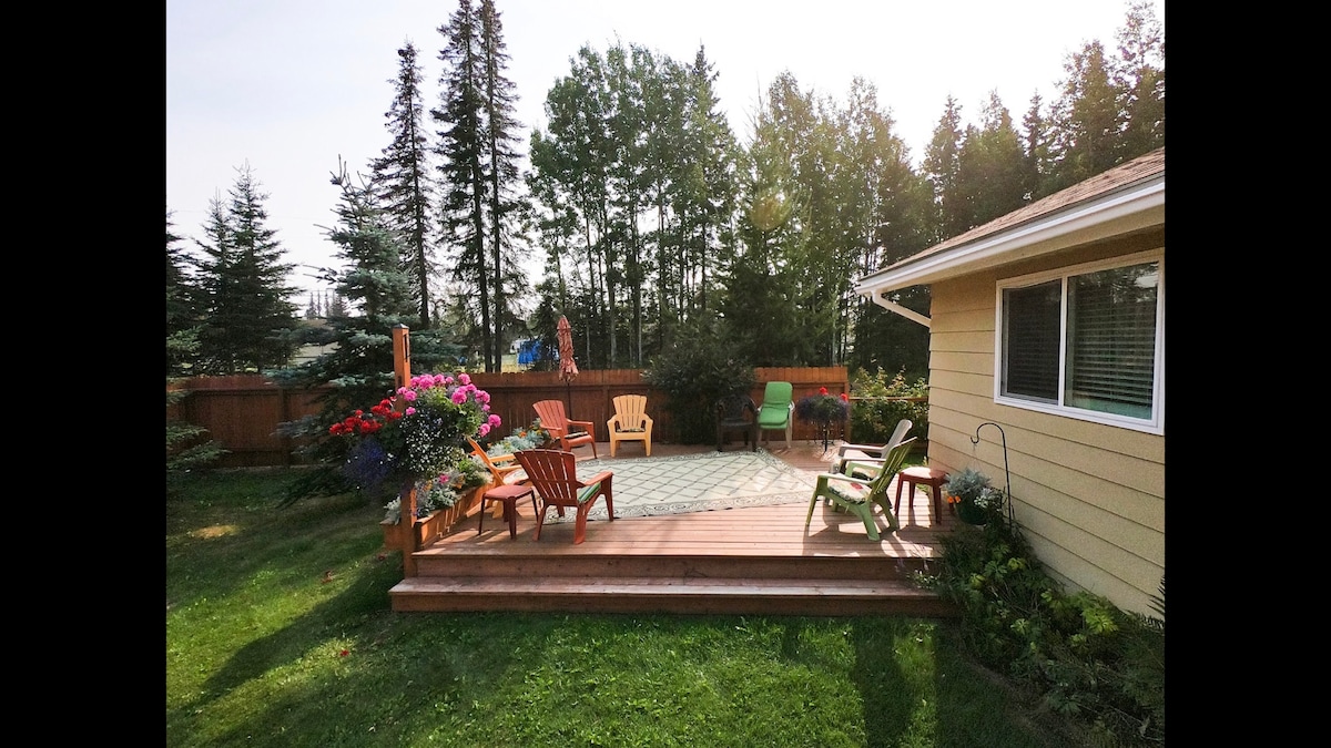 A wooden deck is presented, featuring several colorful Adirondack chairs. The deck is adorned with blooming flower pots and is surrounded by lush greenery and tall trees. A spacious lawn can be seen beyond the deck, offering a serene outdoor setting.