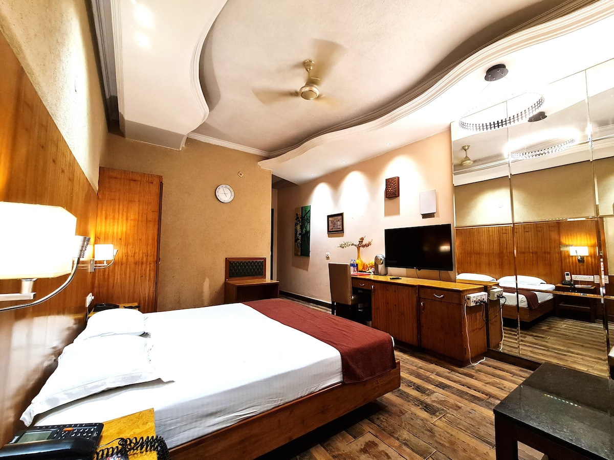 A spacious hotel room features a comfortable bed with crisp white linens and a deep brown blanket. Wood accents are present throughout, including a stylish desk and mirrored wardrobe. Soft lighting creates a relaxed atmosphere, complemented by decorative art pieces on the walls.