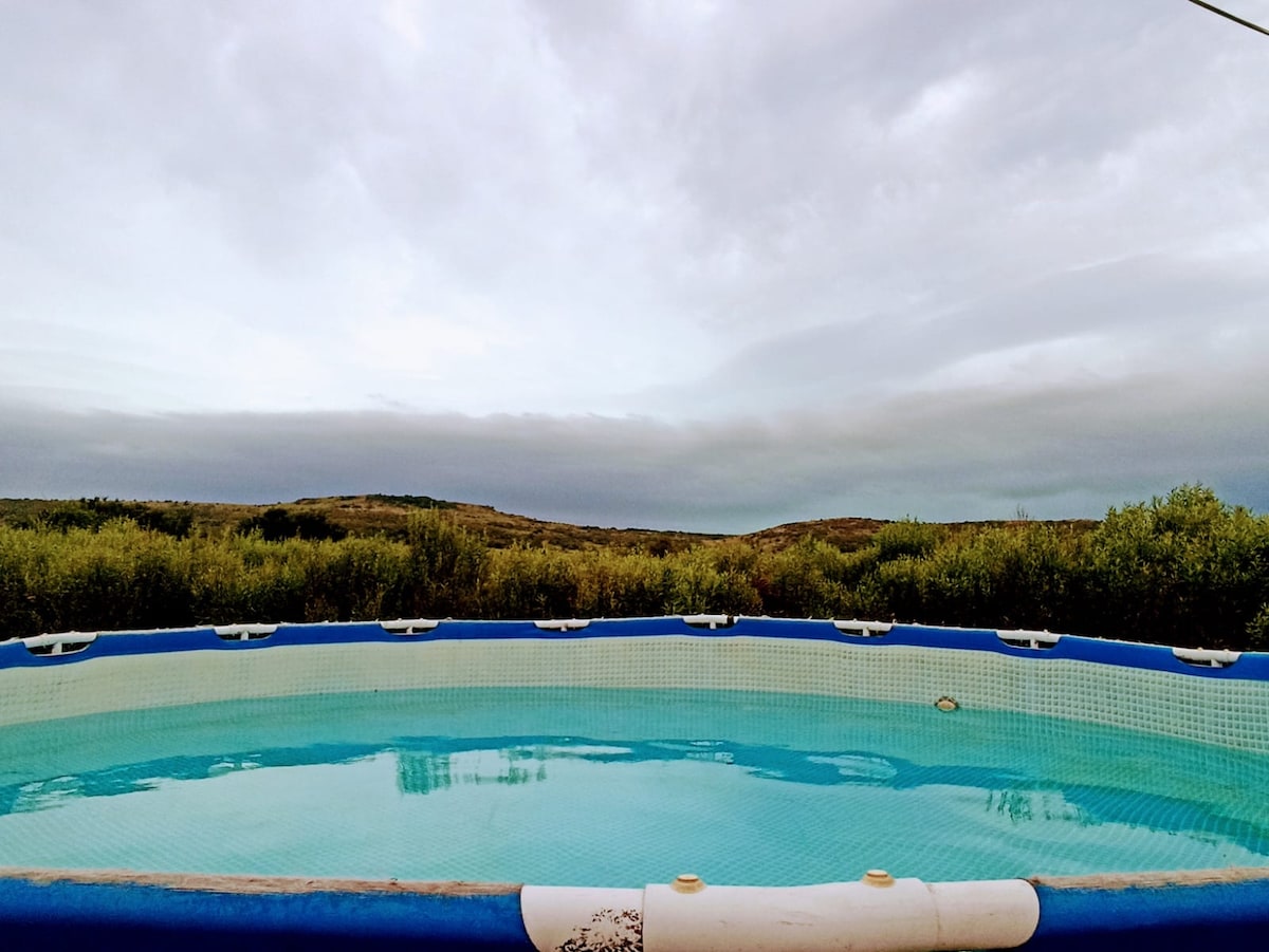 An above-ground pool is visible, surrounded by natural vegetation and rolling hills in the background. The water appears calm and clear, reflecting the overcast sky, creating a serene outdoor setting ideal for relaxation.