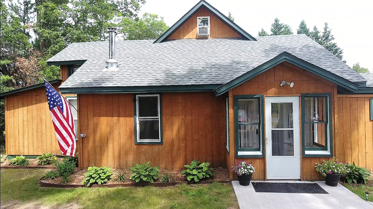 A rustic wooden exterior is showcased, featuring a sloped roof and a welcoming front porch. Brightly colored flower pots adorn the entrance, while a flag is displayed, adding a touch of tradition. Lush green landscaping surrounds the home, enhancing its outdoor appeal.