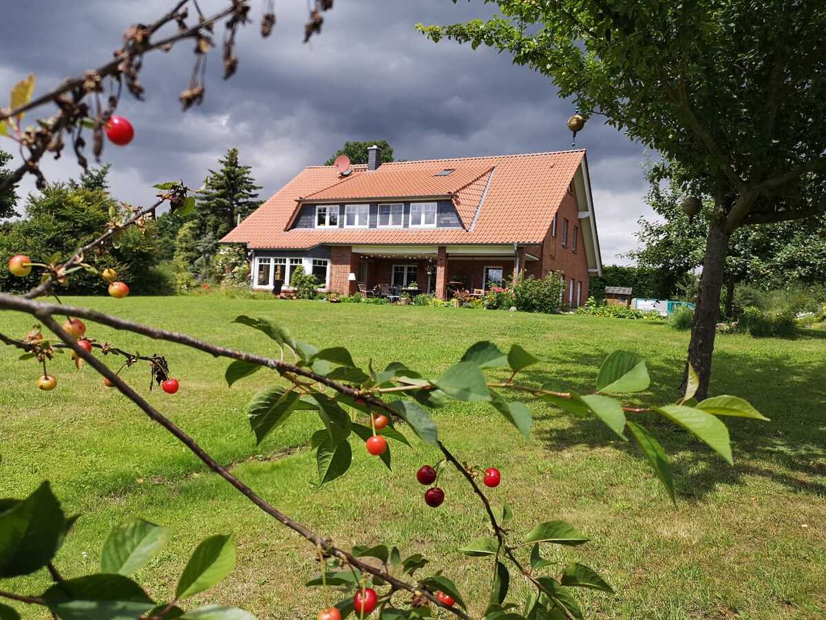 A two-story brick house is framed by a spacious green lawn and trees. The home features large windows and a sloped roof, with a welcoming porch. Berry-laden branches in the foreground enhance the natural setting under a clouded sky.