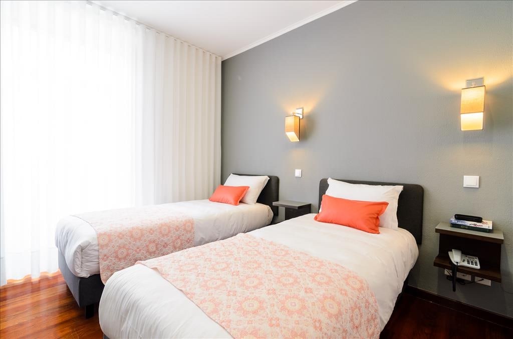 The room contains three single beds, each dressed in white linens with decorative coral pillows. Soft lighting is provided by wall sconces, and a bedside table is positioned next to each bed. Sheer curtains allow natural light to enter from a large window.