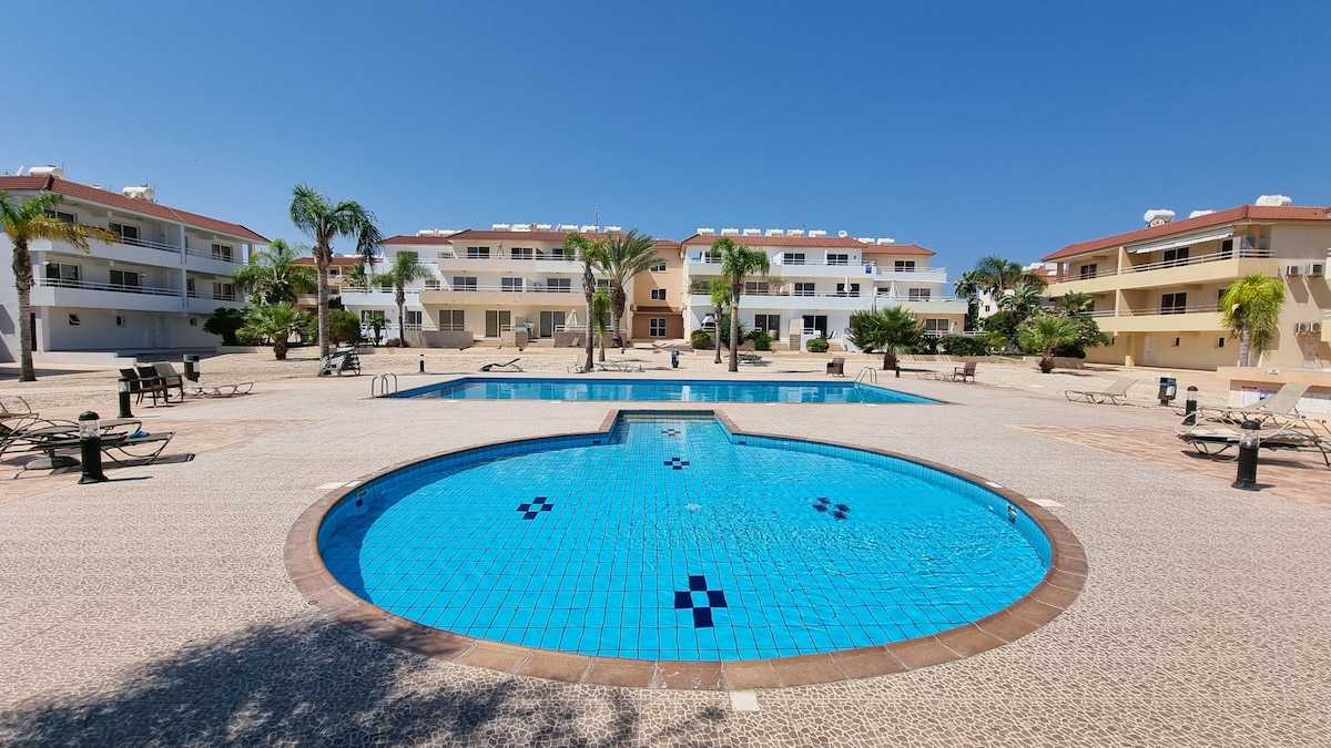 The image captures a large swimming pool area within a complex, featuring a main rectangular pool and a smaller circular pool. Surrounding areas include palm trees and lounge chairs on a textured surface. Bright blue skies and well-maintained buildings are visible in the background.