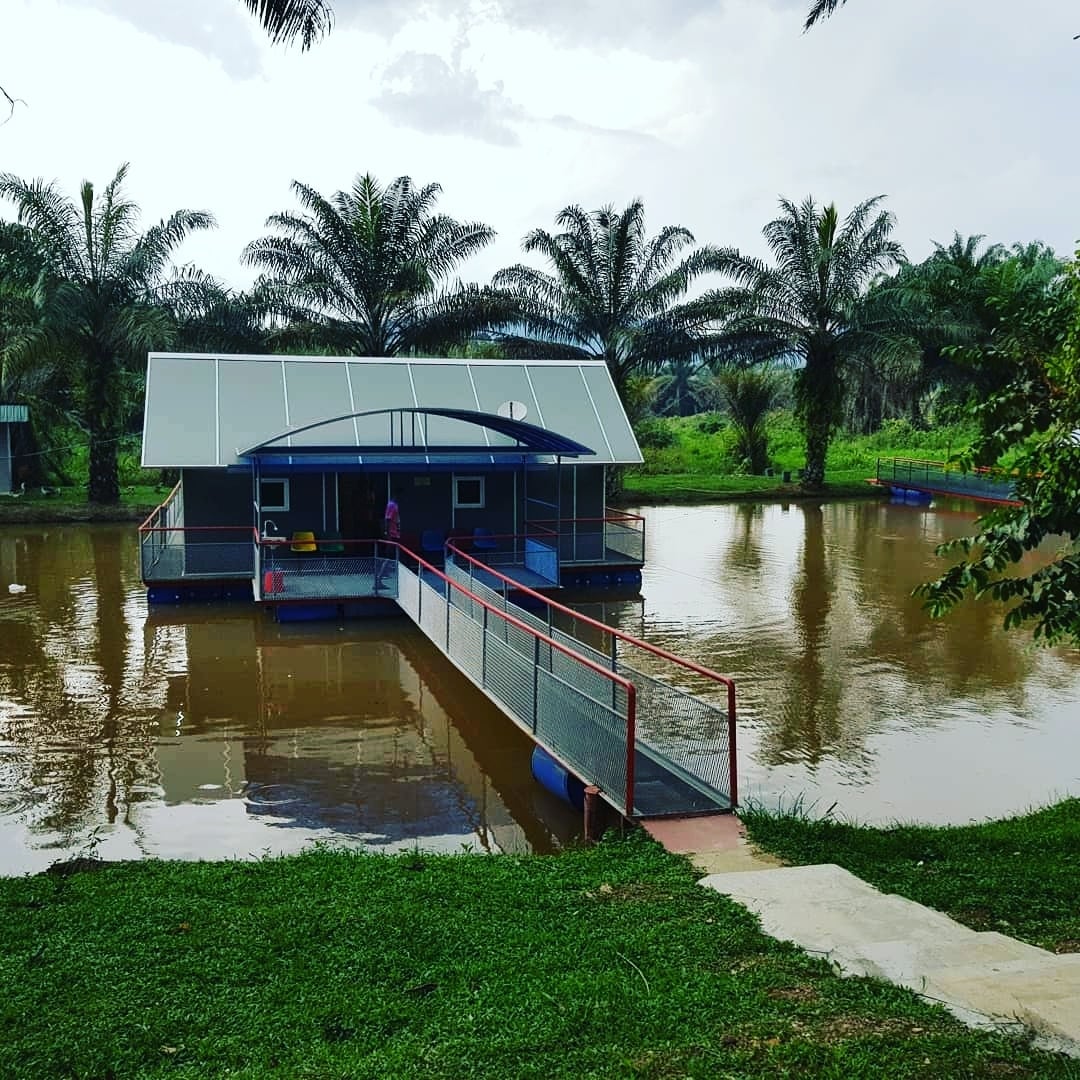A tranquil floating house is visible on a calm lake, surrounded by lush green palm trees. A walkway leads from the grassy shore to the house, creating a welcoming entry point for guests. The structure features a contemporary roof and wide windows, providing views of nature.