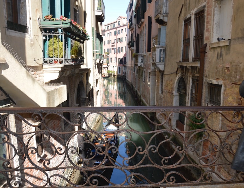 A charming canal is viewed from a decorative iron bridge, flanked by buildings with balconies adorned with potted plants. Reflections of the surrounding architecture are visible on the calm water, enhancing the serene atmosphere of the narrow waterway.