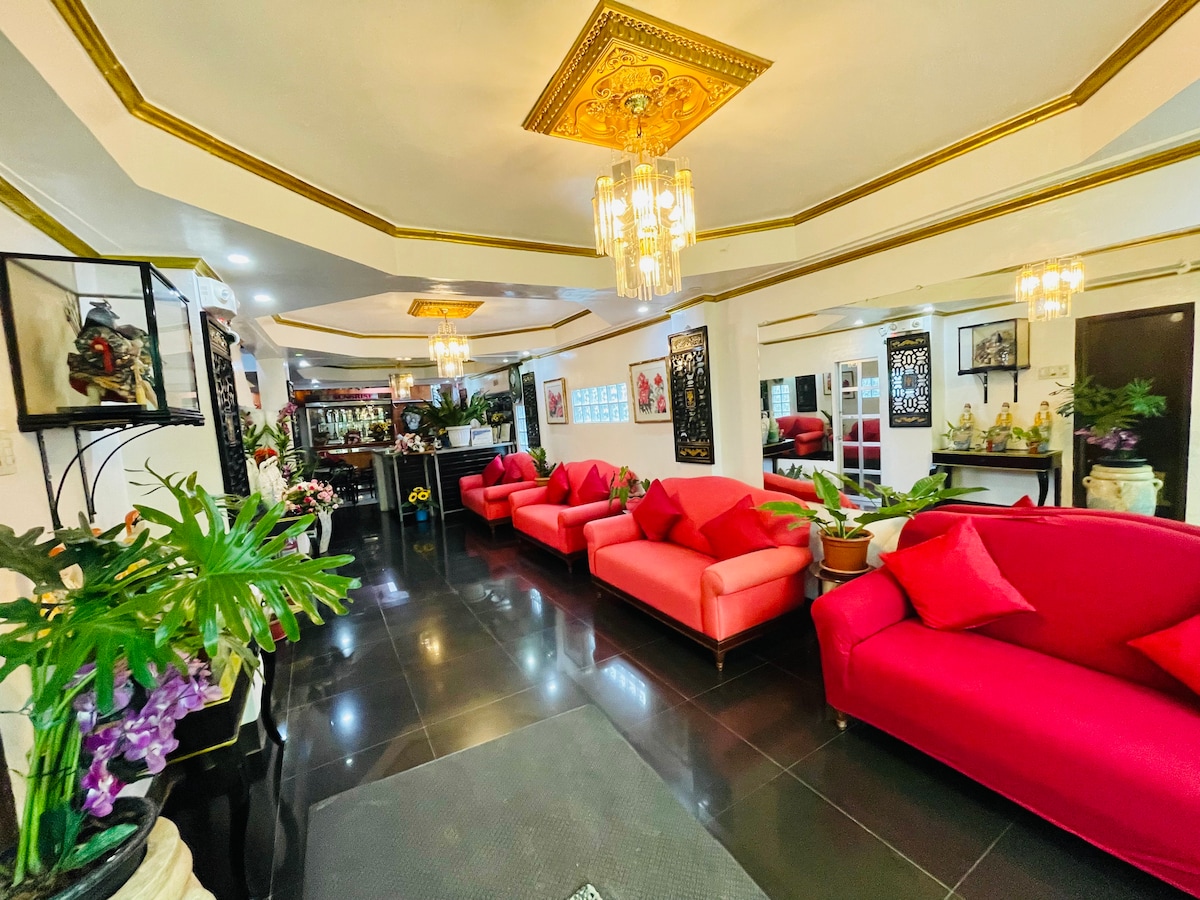 A spacious lobby is featured with vibrant red sofas arranged around a dark tiled floor. Decorative plants and wall art add to the ambiance, while elegant lighting fixtures hang overhead, providing a warm glow. Mirrors reflect the room's design and enhance the feeling of openness.