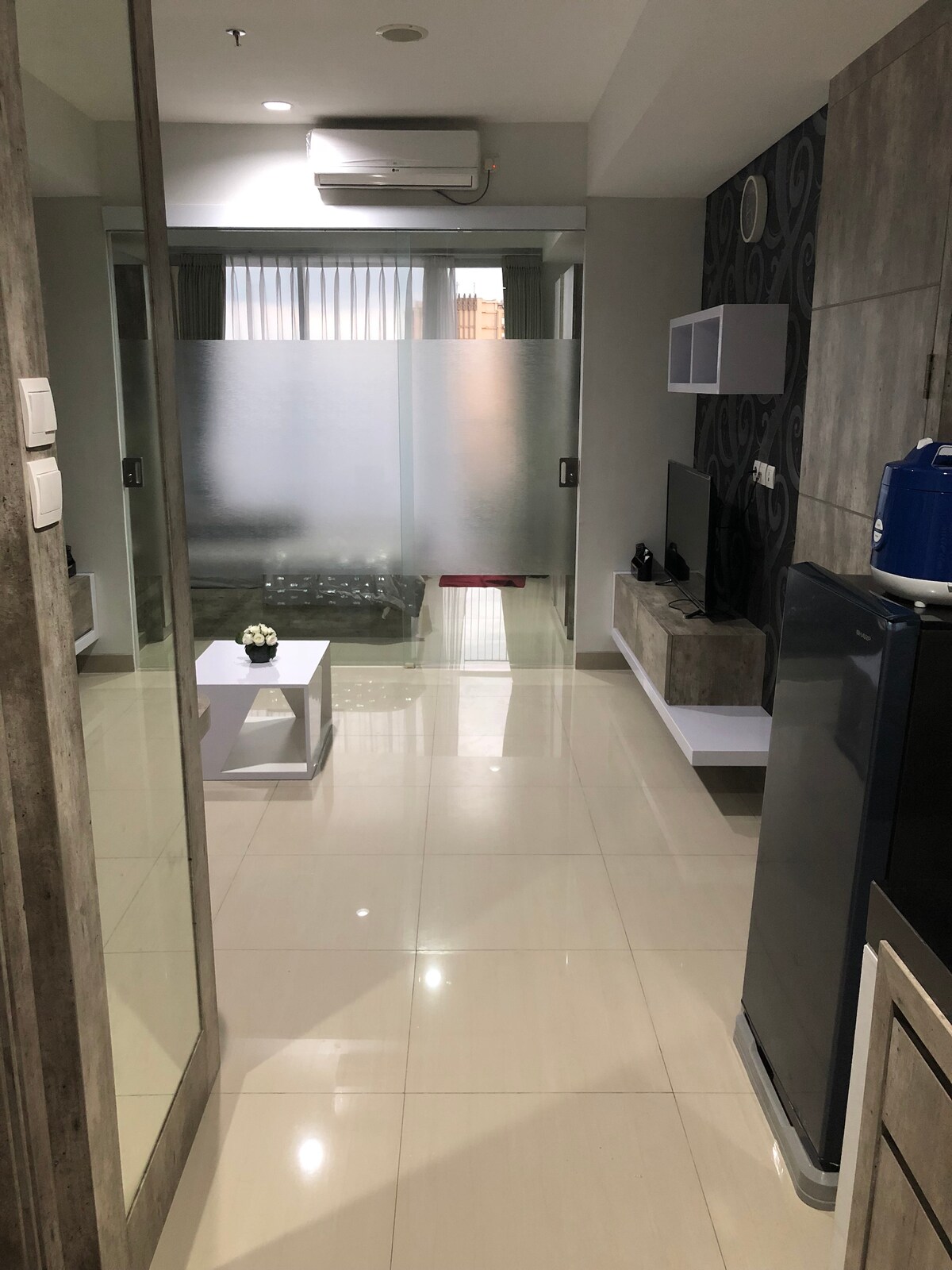 A bright hallway connects various areas within the apartment. Frosted glass panels provide a glimpse into the living space, where a sofa and TV are visible. The tiles reflect light, enhancing the spacious feeling. A small decorative plant rests on a white table.