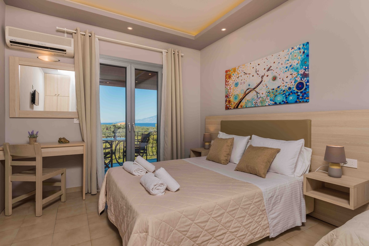 A spacious guest room features two single beds topped with neutral bedding. A wooden desk and chair occupy one corner, while a mirror reflects natural light. Double doors open to a balcony with a pleasant view, and colorful artwork decorates the wall.