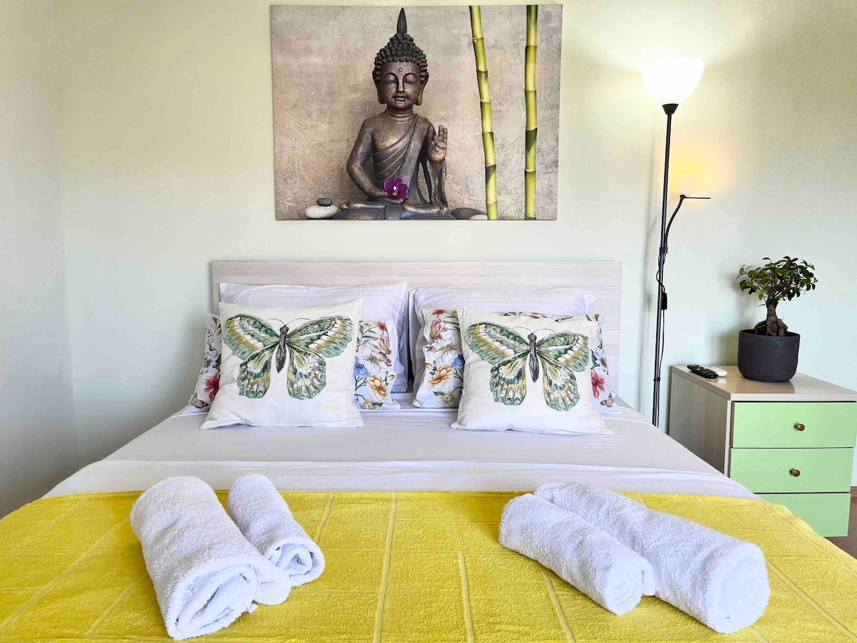 A cozy sleeping area is highlighted by a spacious bed adorned with butterfly-patterned pillows. Above the bed, a serene Buddha artwork enhances the ambiance. A small plant is positioned next to the bed, complemented by a bright yellow blanket and neatly rolled towels.