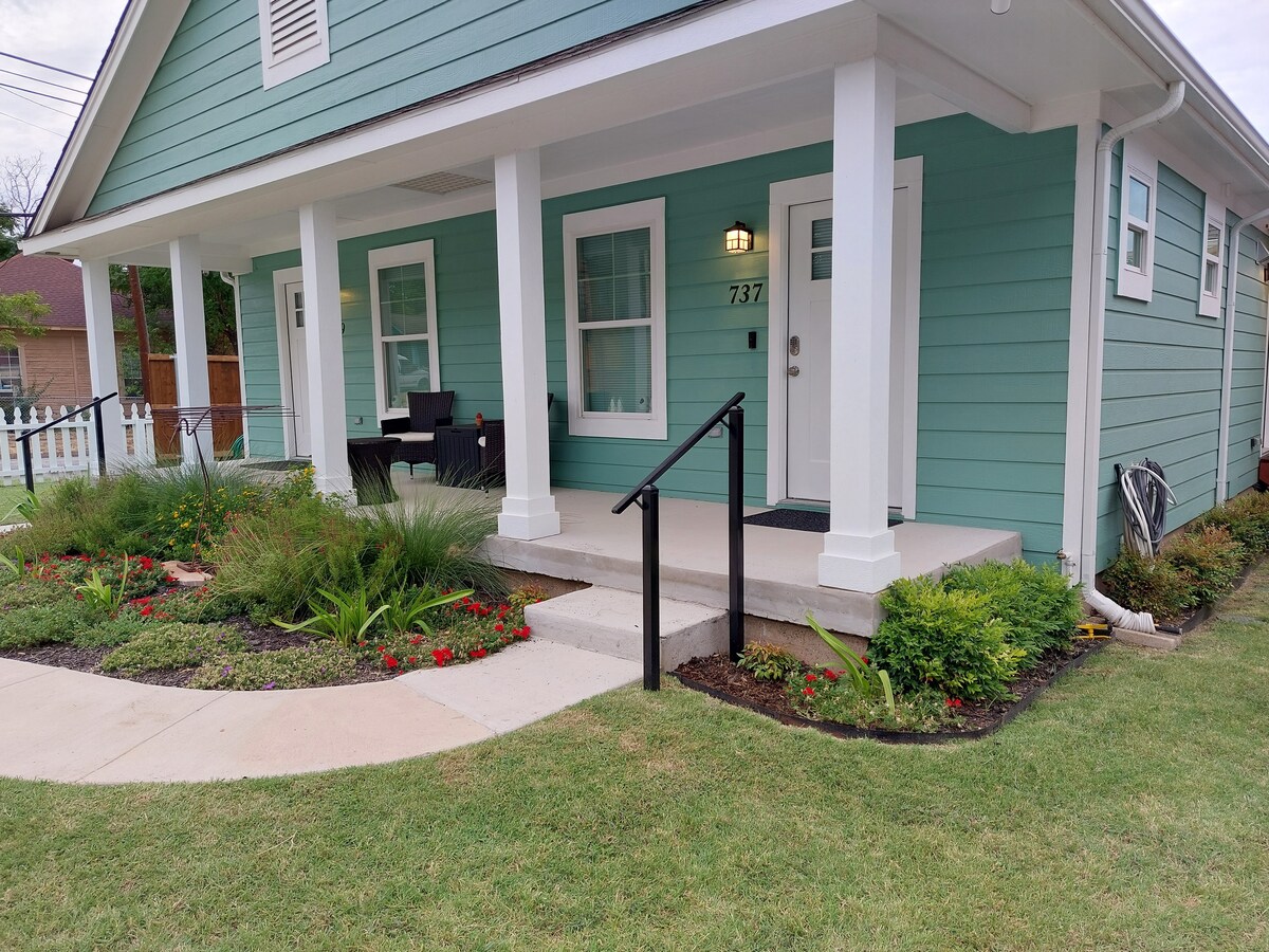 The exterior of the duplex features a light blue facade with white columns and a covered porch. Lush greenery and colorful flower beds enhance the entrance. A paved walkway leads to the front door, with railings for support on the steps.