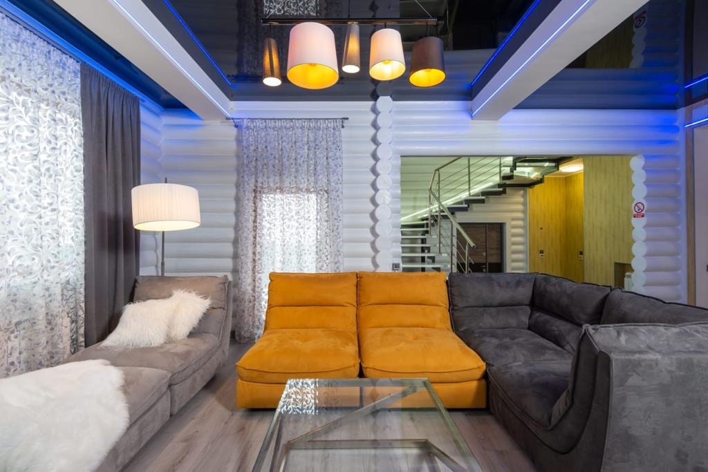 A modern living space features a combination of gray and yellow couches arranged around a glass coffee table. Soft lighting is provided by multiple lamps, and sheer curtains frame the windows, allowing natural light to enhance the setting.
