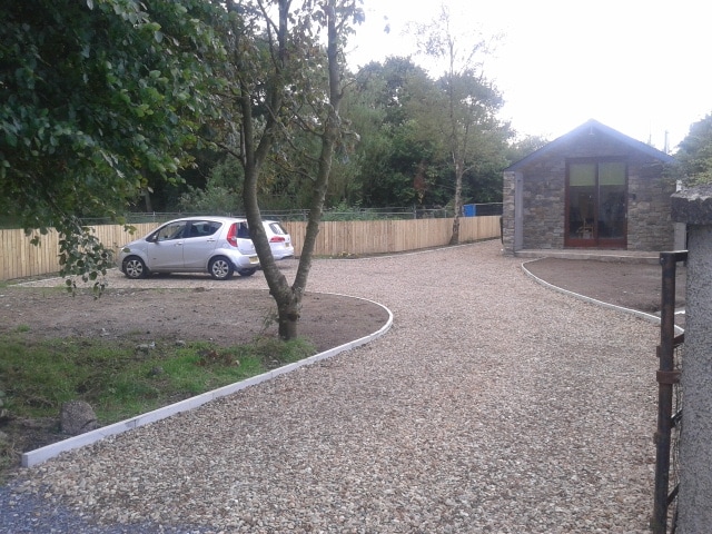 A gravel driveway provides access to a spacious area with parked vehicles. Surrounding trees and a wooden fence frame the property, enhancing privacy. The stone building with large glass doors is visible in the background, indicating a welcoming entryway.