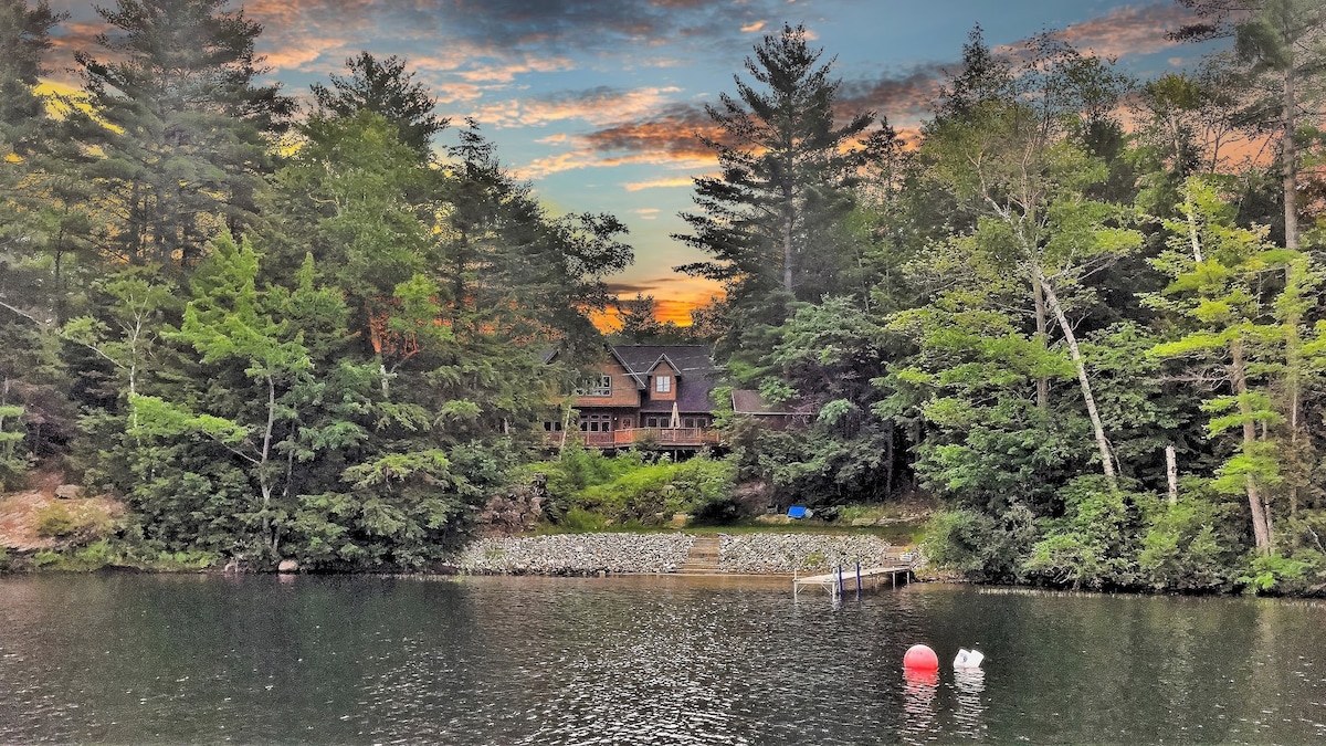 A serene lakefront home is nestled among lush trees, with a backdrop of a colorful sunset. The calm water reflects the vibrant hues of the sky. A small dock extends into the pond, bordered by natural landscaping.