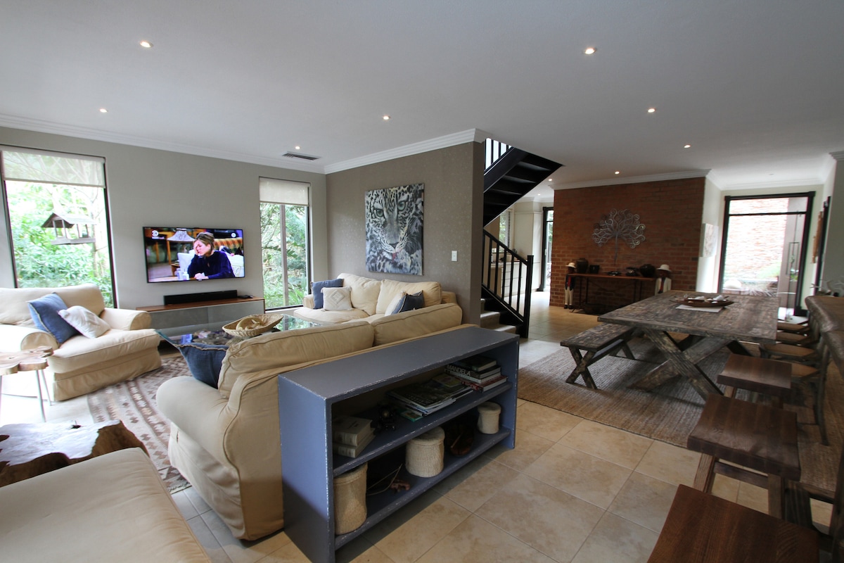 A spacious living area is showcased with light-colored sofas arranged around a coffee table. A large TV is mounted on the wall, reflecting bright indoor lighting. Dining tables with wooden chairs can be seen in the background, complemented by large windows allowing natural light to fill the space.