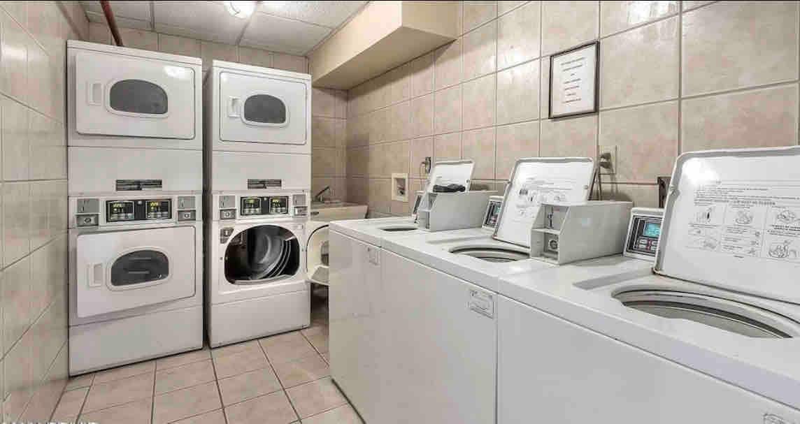 A clean and organized laundry room is equipped with stacked washer and dryer units as well as a front-loading washing machine. The space is tiled, with bright overhead lighting illuminating the area. Instructional signs are posted for user convenience.