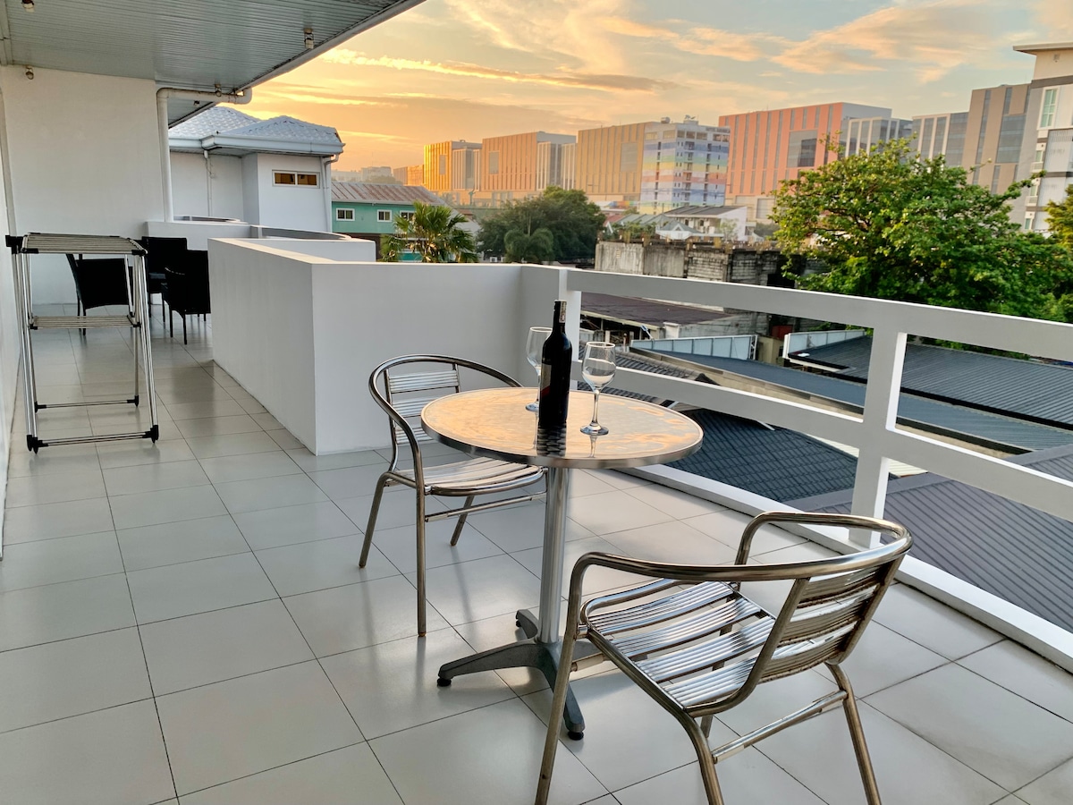 A spacious balcony features a table with two metallic chairs, inviting relaxation. A bottle of wine rests on the table, set against a backdrop of a golden sunrise and vibrant city skyline. The area is equipped with tiled flooring, enhancing outdoor comfort.